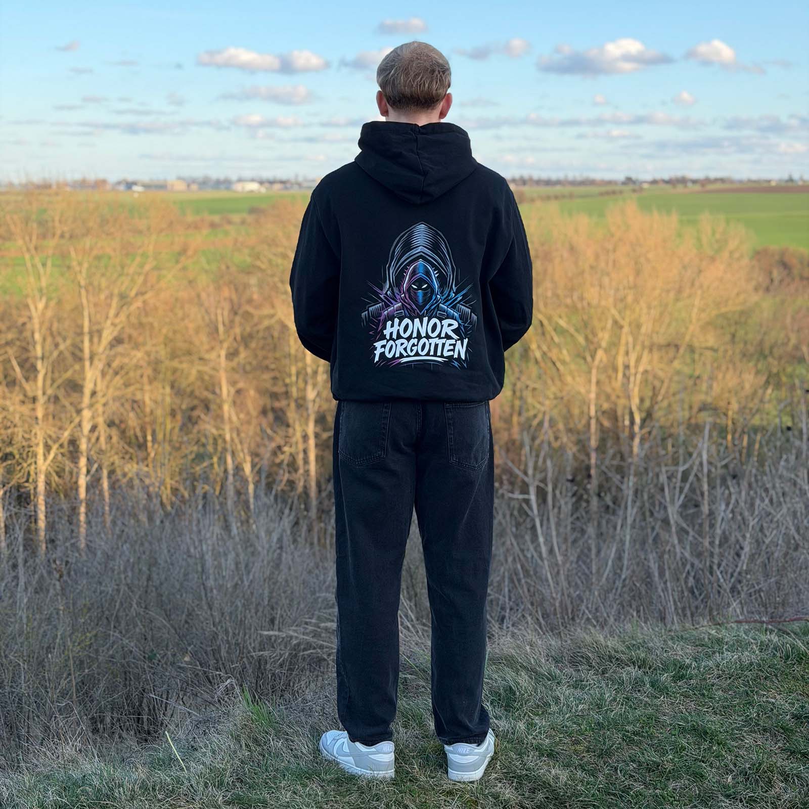 Rückansicht eines schwarzen Honor Forgotten Hoodies am Model im Outdoor-Setting, großer grafischer Backprint sichtbar.
