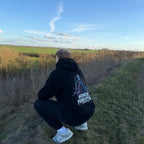 Model trägt schwarzen Honor Forgotten Hoodie in gehockter Pose von hinten, großer Backprint im Outdoor-Setting sichtbar.