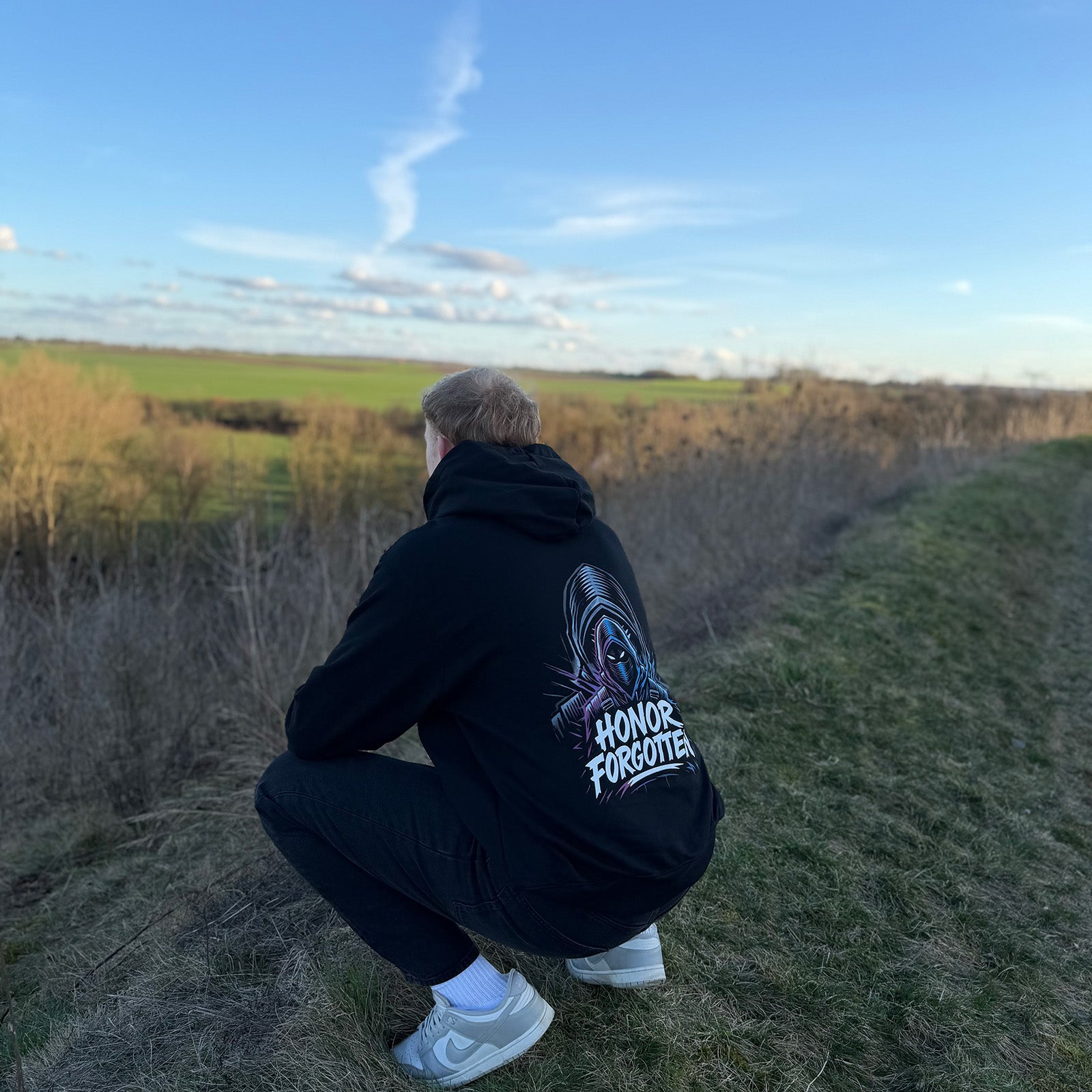 Model trägt schwarzen Honor Forgotten Hoodie in gehockter Pose von hinten, großer Backprint im Outdoor-Setting sichtbar.